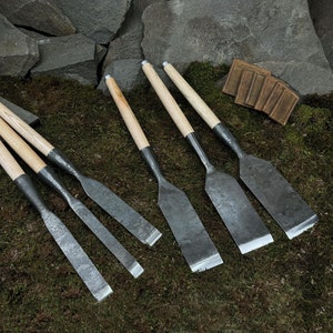 Puede incluir: Conjunto de seis cinceles de metal con mango de madera y diferentes longitudes de hoja, dispuestos sobre un lecho de musgo verde.