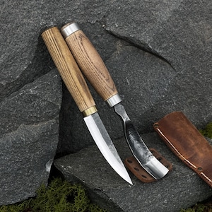 May include: Two wood-handled carving knives with silver blades rest on a gray stone surface. One knife has a straight blade, the other a curved blade. A brown leather sheath is next to the knives.