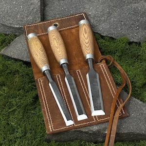 May include: A set of three wood chisels with light brown wooden handles and silver metal blades, arranged inside a brown leather tool roll with white stitching and a long leather tie strap for securing the set when it is rolled up.