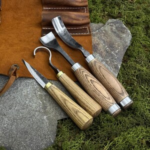 May include: A set of four wood carving tools with wooden handles and metal blades. The tools are arranged on a brown leather roll-up case. The blades have different shapes, including a knife, a hook, and a curved gouge.