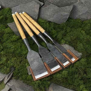 Könnte beinhalten: Ein Set aus vier Holzmeißeln mit langen, hellen Holzgriffen mit sichtbarer Maserung. Jede Metallklinge ist dunkel, geschmiedet und flach, mit einer scharfen, polierten Schneide. Jeder Meißel ist durch eine braune Lederscheide geschützt.