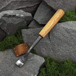May include: A hand tool featuring a dark, forged metal blade with a curved, sharpened edge and a long, cylindrical wooden handle with a visible grain. A small, square, tan leather sheath with four metal rivets sits beside the metal tool.