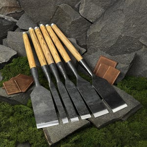 May include: A set of five wood chisels with dark, forged metal blades and light-coloured wooden handles. The tools are arranged in a row, featuring varying blade widths, with metallic ferrules connecting the steel to the wooden grips.