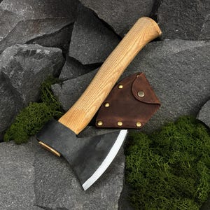 Può includere: Un'accetta con manico in legno chiaro e testa in metallo forgiato scuro con lama affilata. Un fodero in pelle marrone con rivetti in metallo color ottone e chiusura a scatto è posizionato accanto alla testa dell'accetta.