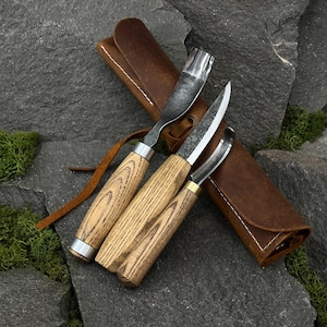 May include: Three wood carving tools with light-coloured wood handles and metal blades, including a curved gouge, a straight blade, and a smaller hook knife. The tools are arranged on top of a brown leather roll-up case with a leather tie closure.