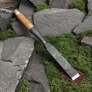 Puede incluir: Un cincel de acero forjado a mano con una hoja de metal larga, plana y de color gris oscuro con un borde biselado y un mango cilíndrico de madera con una virola de metal plateado en la parte superior, apoyado sobre un trozo de cuero marrón.