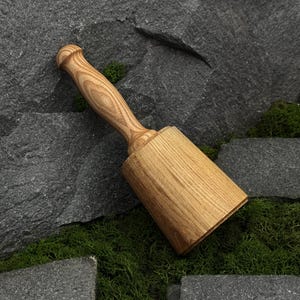 May include: A wooden mallet featuring a cylindrical head and a tapered handle with a visible natural grain pattern. The tool is crafted from light brown wood, showcasing a smooth, polished finish throughout its entire structure.