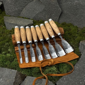 May include: A collection of eight wood carving chisels with wooden handles and metal blades, presented in a brown leather tool roll. The chisels are arranged on green moss, with large grey rocks in the background.