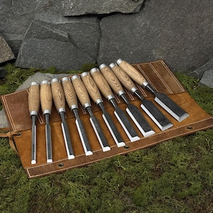 May include: A set of ten wood chisels with light brown wooden handles and silver metal ferrules, arranged in a row on a brown leather tool roll. Each chisel features a flat, sharp steel blade with a beveled edge, secured in a dedicated leather slot.