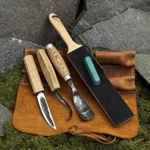 May include: A wood carving set with three knives and a sharpening tool. The knives have wooden handles and metal blades. The sharpening tool has a black surface and a green sharpening stone. The set is in a brown leather pouch.