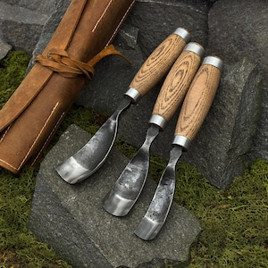 May include: Three wood carving gouges with wooden handles and metal blades are arranged on a dark stone surface. A brown leather tool roll is in the background, suggesting a craft or woodworking theme.