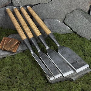 Pode incluir: Quatro formões para madeira com cabos de madeira castanho-claro e lâminas de metal escuro e texturizado. Os formões estão dispostos lado a lado, apresentando bordas afiadas e chanfradas nas pontas. Uma pequena bainha de couro castanho está próxima.
