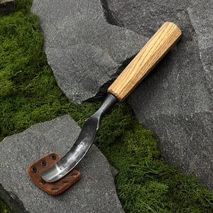 Puede incluir: Una herramienta para tallar madera con una hoja de metal curva de acabado pulido y un mango de madera de color marrón claro. La hoja está protegida por una pequeña funda cuadrada de cuero marrón asegurada con cuatro remaches metálicos.