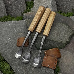 Puede incluir: Tres herramientas de metal forjadas a mano con mangos de madera clara y hojas anchas y curvas. Cada herramienta presenta un eje de metal gris oscuro texturizado y un borde pulido. Se incluyen dos pequeños protectores de cuero marrón con remaches metálicos.