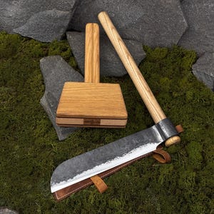Könnte beinhalten: Ein Holzhammer und ein großes, gebogenes Messer mit einer Lederscheide werden auf grünem Moos präsentiert. Das Messer hat eine dunkle, strukturierte Klinge und einen Holzgriff. Der Hammer ist aus hellem Holz gefertigt.