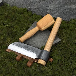May include: A handcrafted, rustic-style cleaver with a wooden handle and a leather sheath. A wooden mallet is also present. The blade has a textured finish. The items are displayed on a bed of green moss.