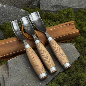 Può includere: Tre sgorbie per intaglio del legno con lame in metallo e manici in legno. I manici presentano un motivo a venature del legno marrone chiaro e sono accentuati da anelli in metallo color argento. Un astuccio per attrezzi in pelle marrone è sullo sfondo, appoggiato su una superficie di pietra.