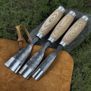 May include: Four wood carving chisels with light brown wooden handles and silver metal accents. The blades are made of dark metal and are arranged on a brown leather surface. The background is a mix of green moss and a gray stone.