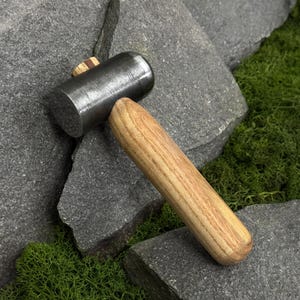 May include: A mallet featuring a cylindrical, dark grey metal head with a visible wood grain wedge insert and a smooth, light brown wooden handle. The tool is positioned at an angle, showcasing the contrast between the metallic and wooden parts.