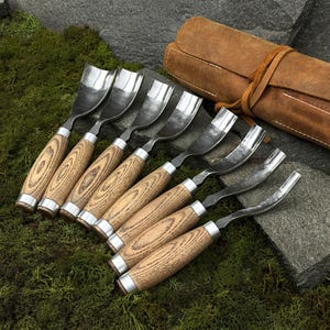 Peut inclure: Un ensemble de huit gouges à bois avec des lames en acier poli et des manches en bois. Les manches présentent un motif de grain de bois brun clair et des viroles en métal argenté. Un rouleau d'outils en cuir marron est visible en arrière-plan.