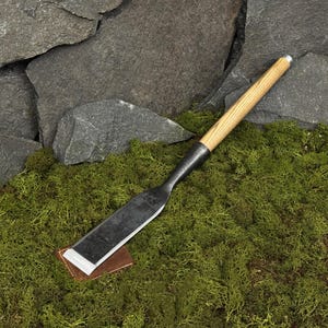 May include: A hand-forged chisel with a wooden handle and a dark metal blade. The chisel rests on a small, rectangular copper plate, set against a backdrop of green moss and gray stone.