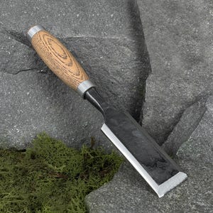 Peut inclure: Un ciseau à bois doté d'une lame en métal forgé sombre avec un bord biseauté et un manche cylindrique en bois au grain apparent, fixé par une virole en métal argenté lisse à la base du manche.
