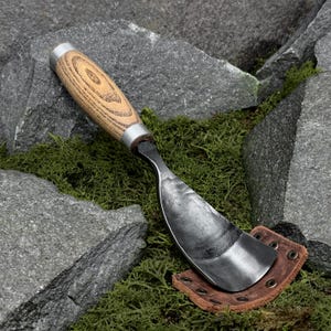 Op de afbeelding: Een handgesmeed metalen tuinschepje met een gebogen, donkergrijs stalen blad met een gehamerde textuur, een lichtbruin houten handvat met een zilverkleurige metalen huls en een kleine bruine lederen schede met metalen klinknagels.