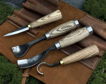 Op de afbeelding: Een set van vier houtsnijgereedschappen met lichtgekleurde houten handvatten en metalen ringen, met diverse bladvormen waaronder een recht mes, een brede guts, een gebogen guts en een haakmes, op een bruine leren rol.