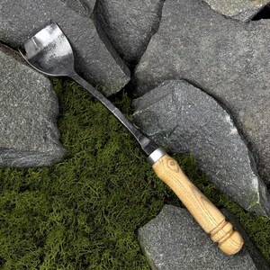 Pode incluir: Uma ferramenta de jardim em metal forjado &agrave; m&atilde;o com uma cabe&ccedil;a larga e curva de acabamento escuro e texturizado, e um cabo longo de madeira clara. O cabo tem uma forma cil&iacute;ndrica lisa com an&eacute;is decorativos ranhurados na base.