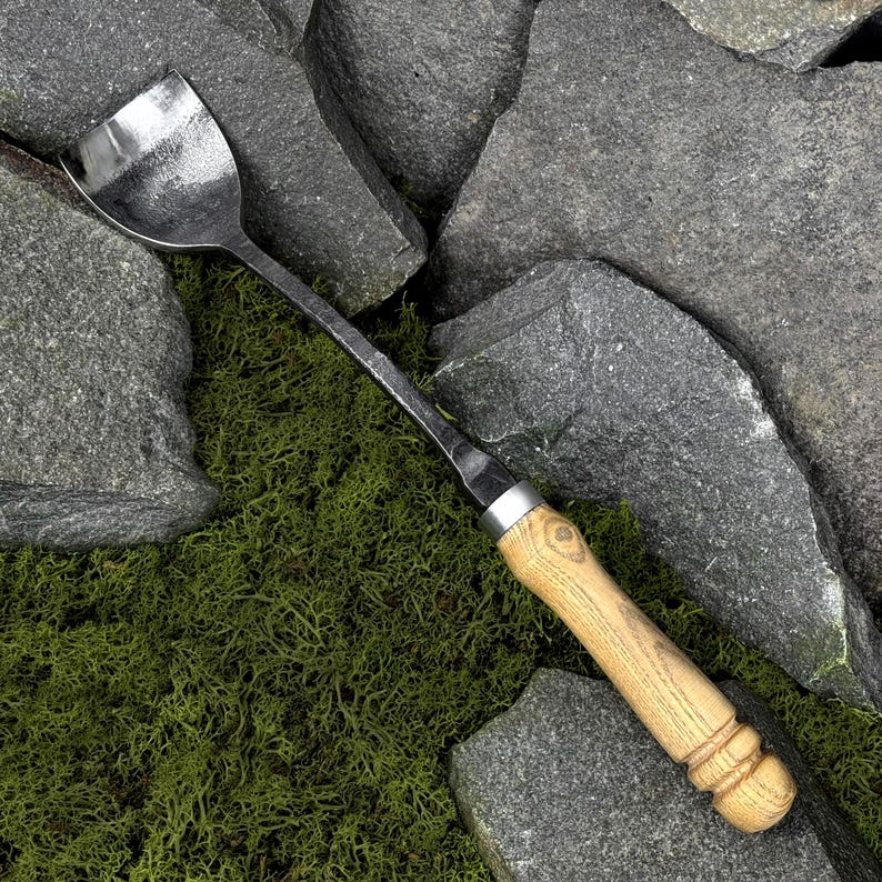 Pode incluir: Uma ferramenta de jardim em metal forjado &agrave; m&atilde;o com uma cabe&ccedil;a larga e curva de acabamento escuro e texturizado, e um cabo longo de madeira clara. O cabo tem uma forma cil&iacute;ndrica lisa com an&eacute;is decorativos ranhurados na base.