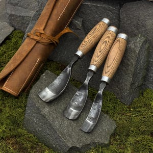 May include: Three wood carving gouges with curved metal blades and light brown wooden handles, accompanied by a brown leather tool roll. Each tool features a silver-colored metal ferrule connecting the handle to the forged steel blade.