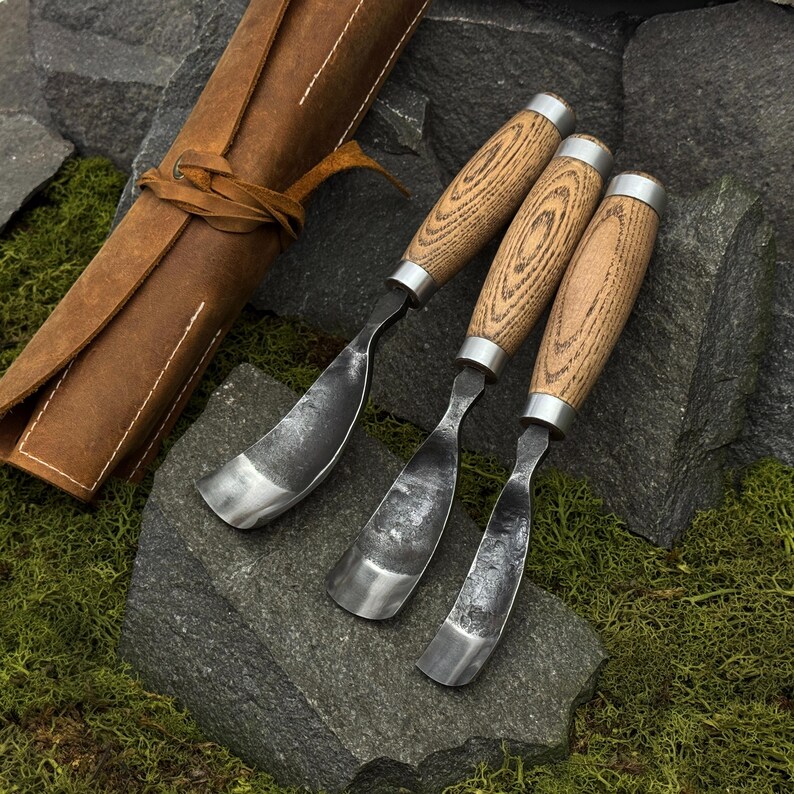 May include: Three wood carving gouges with curved metal blades and light brown wooden handles, accompanied by a brown leather tool roll. Each tool features a silver-colored metal ferrule connecting the handle to the forged steel blade.