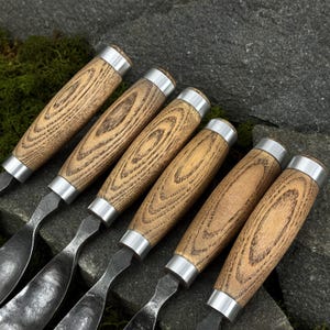 May include: A set of six hand tools featuring textured, dark-toned metal blades and cylindrical wooden handles with visible grain patterns. Each handle is secured with a polished silver-colored metal ferrule at the top, creating a uniform appearance.