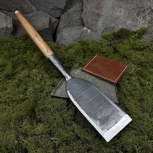 May include: A long-handled, flat-bladed tool with a wooden handle and a metal blade. The blade is slightly curved and has a sharp edge. The tool is resting on a bed of moss and rocks.