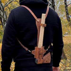 May include: A brown leather axe carrier with adjustable straps and a buckle closure. The carrier holds a wooden-handled axe, and is designed to be worn on the back. The background shows a forest with yellow leaves.