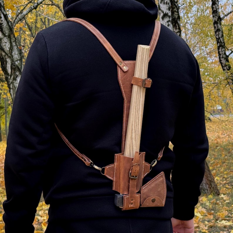 May include: A brown leather axe carrier with adjustable straps and a buckle closure. The carrier holds a wooden-handled axe, and is designed to be worn on the back. The background shows a forest with yellow leaves.
