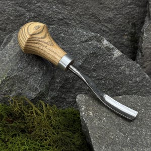 Puede incluir: Una gubia para tallar madera con una hoja curva de color plateado y un mango de madera marrón claro. El mango tiene forma redondeada con una virola de metal plateado. La herramienta está diseñada para carpintería y tallado.