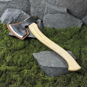 Peut inclure: Hache avec un manche en bois clair et une tête en métal foncé. La tête de la hache a une lame argentée et un étui en cuir marron. La hache repose sur un lit de mousse verte et de roches grises.