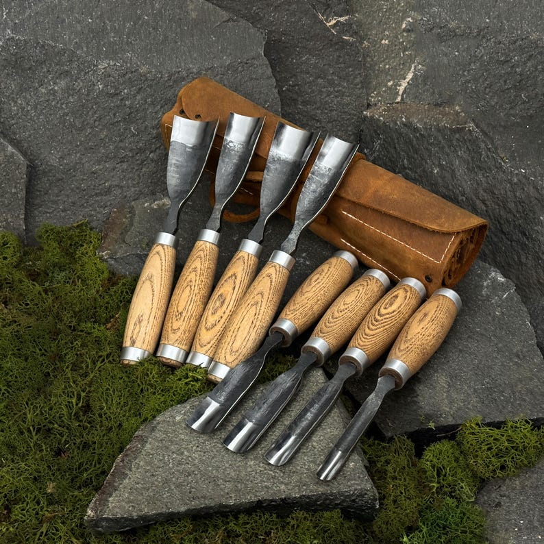 May include: Set of wood carving chisels with light wood handles and metal blades. The chisels are arranged on a dark stone surface with a leather tool roll. The handles have a natural wood grain pattern.