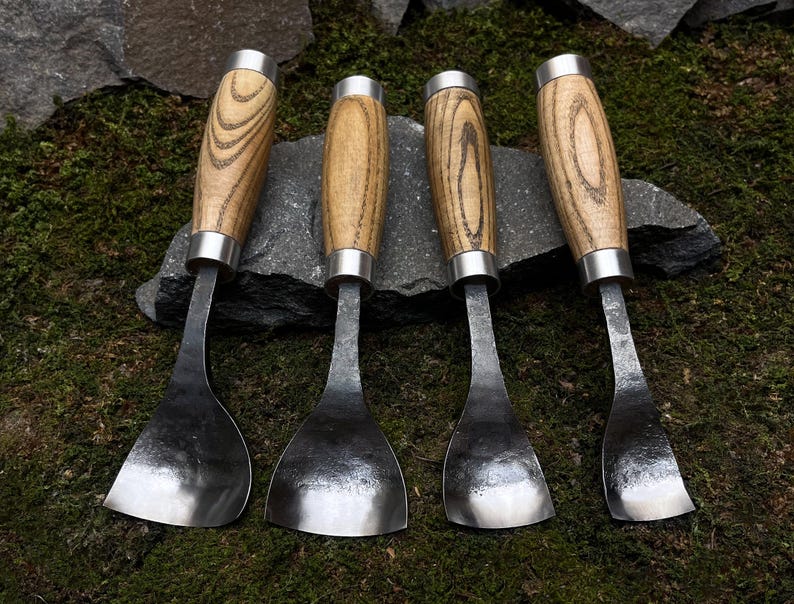 May include: Four wood-handled carving tools with curved metal blades. The tools are arranged in a row on a bed of moss and rocks.