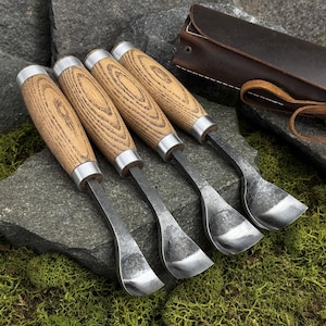 May include: Four hook knives with light brown wooden handles and silver metal ferrules. The blades are curved, dark, and textured, featuring a spoon-like shape. A dark brown leather roll-up case with a strap is positioned behind the tools.