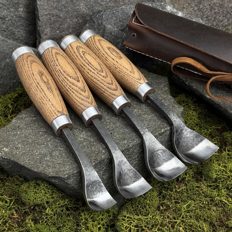May include: Four hook knives with light brown wooden handles and silver metal ferrules. The blades are curved, dark, and textured, featuring a spoon-like shape. A dark brown leather roll-up case with a strap is positioned behind the tools.