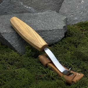 May include: A wood-handled carving knife with a curved blade and a leather sheath. The knife has a light brown wooden handle and a shiny silver blade. The sheath is brown and has a snap closure. The knife is resting on green moss.