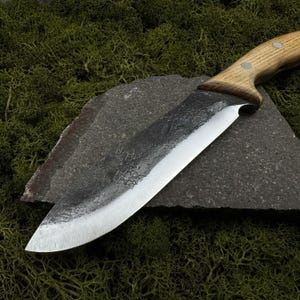 May include: A handcrafted chef's knife with a wooden handle and a dark, textured blade. The blade is made of steel and has a sharp edge. The knife is resting on a dark gray stone, with green moss in the background. A kitchen utensil.
