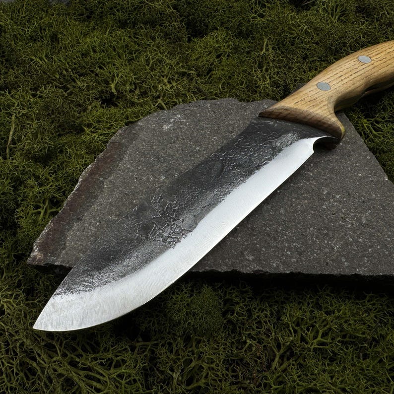 May include: A handcrafted chef's knife with a wooden handle and a dark, textured blade. The blade is made of steel and has a sharp edge. The knife is resting on a dark gray stone, with green moss in the background. A kitchen utensil.