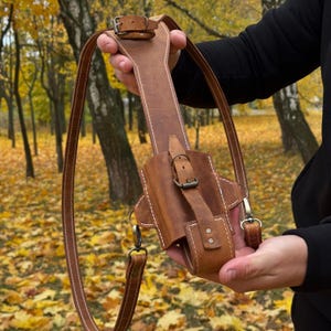 May include: A brown leather holster with a shoulder strap. The holster has a rectangular shape with a curved bottom and is secured with a buckle. The strap is adjustable and has a metal clasp. The stitching is white.