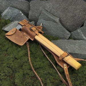 May include: A handcrafted axe with a wooden handle and a leather sheath. The axe head is silver, and the sheath is brown with buckle closures. The axe rests on green moss with a backdrop of gray rocks.