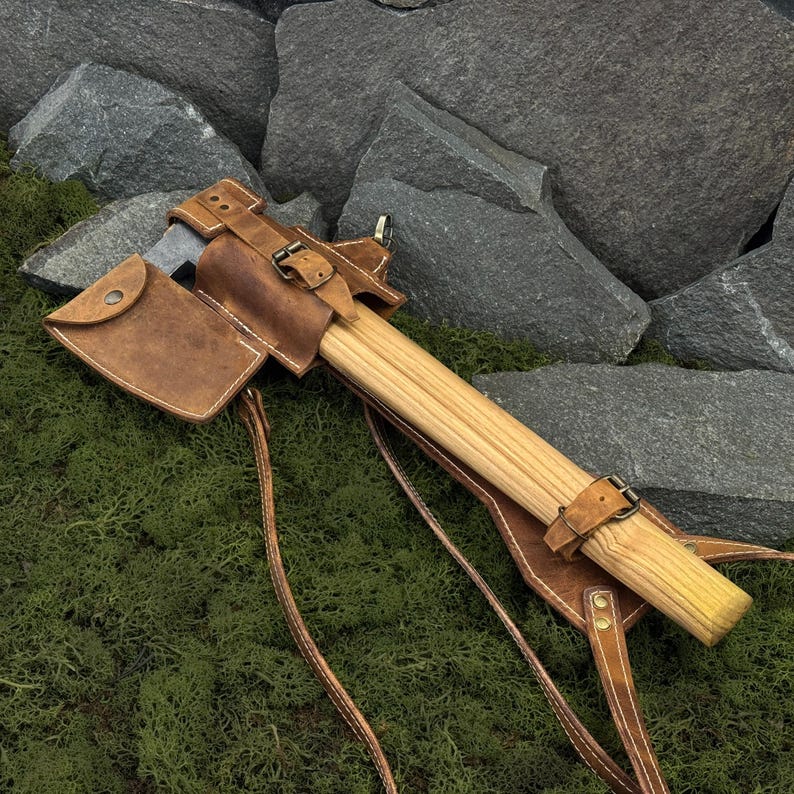 May include: A handcrafted axe with a wooden handle and a leather sheath. The axe head is silver, and the sheath is brown with buckle closures. The axe rests on green moss with a backdrop of gray rocks.