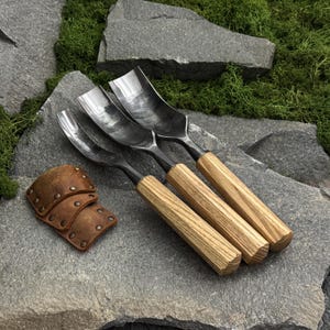 May include: Three metal gouges with curved, dark steel blades and light-colored, hexagonal wooden handles. Beside the tools are three small, rectangular brown leather guards featuring metallic rivets along their edges.