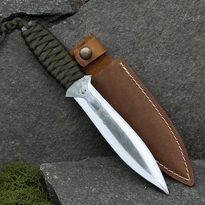 Peut inclure: Un poignard artisanal avec une lame argentée et un manche enveloppé de vert. Le poignard est associé à un étui en cuir marron, fermé par un bouton. La lame mesure environ 20 cm de long.